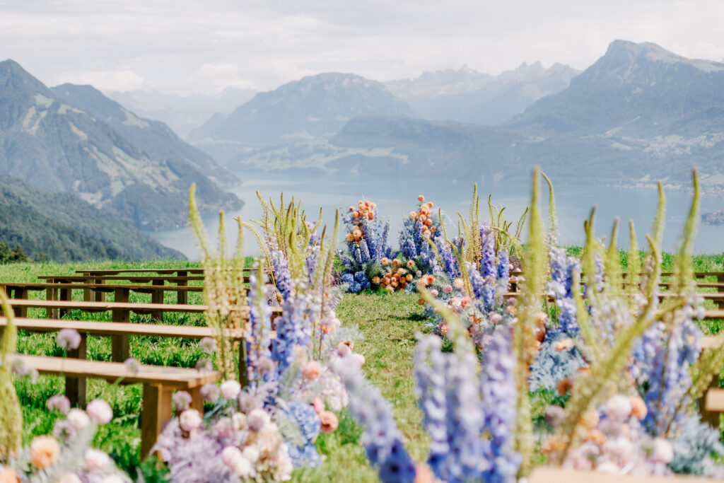 Ein Hochzeitsgang im Freien mit bunten Blumen, Holzbänken und einer malerischen Berg- und Seekulisse, inspiriert von der Schönheit Südtirols.