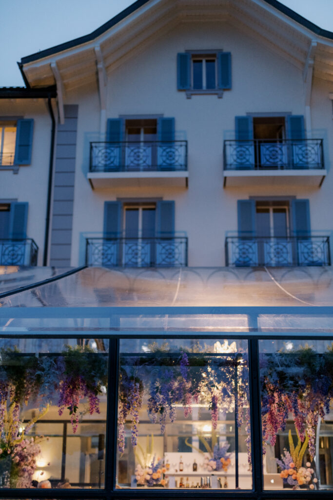 Elegantes Gebäude mit blauen Fensterläden, Balkonen und einem gläsernen Gewächshaus, das mit hängenden Blumen geschmückt ist - perfekt für ein Hochzeitswochenende, das vom Charme Südtirols inspiriert ist.