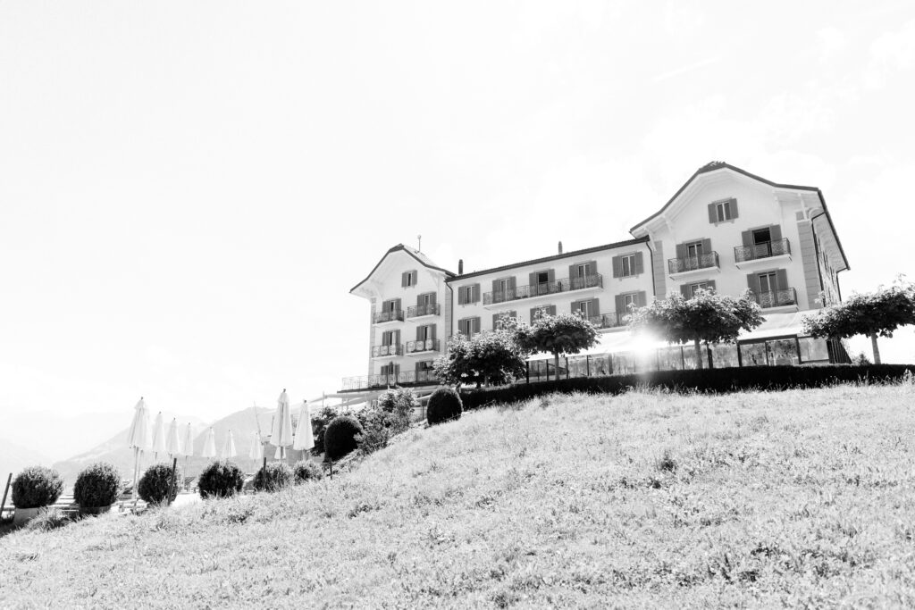 Schwarz-Weiß-Foto eines großen Hotels auf einem grasbewachsenen Hügel in Südtirol, mit Bäumen und Sonnenschirmen auf der Terrasse davor.