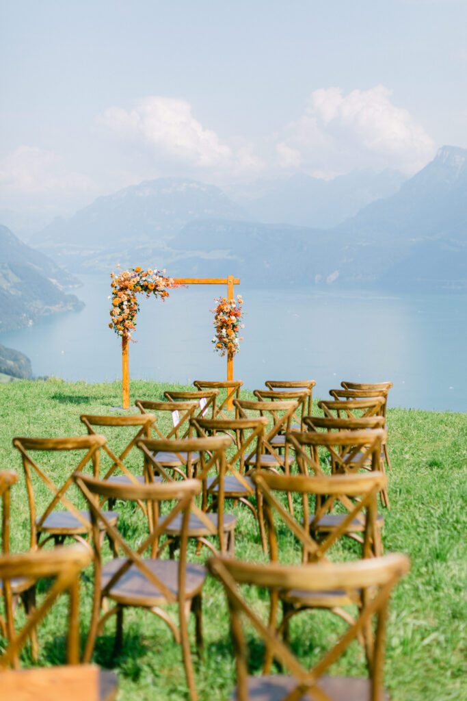 Holzstühle vor einem Blumenbogen im Freien, mit Blick auf einen See und Berge unter blauem Himmel - perfekt für ein Hochzeitswochenende in der Schweiz oder Österreich.